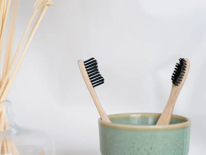 Two bamboo toothbrushes in a pale green ceramic cup against a white wall.