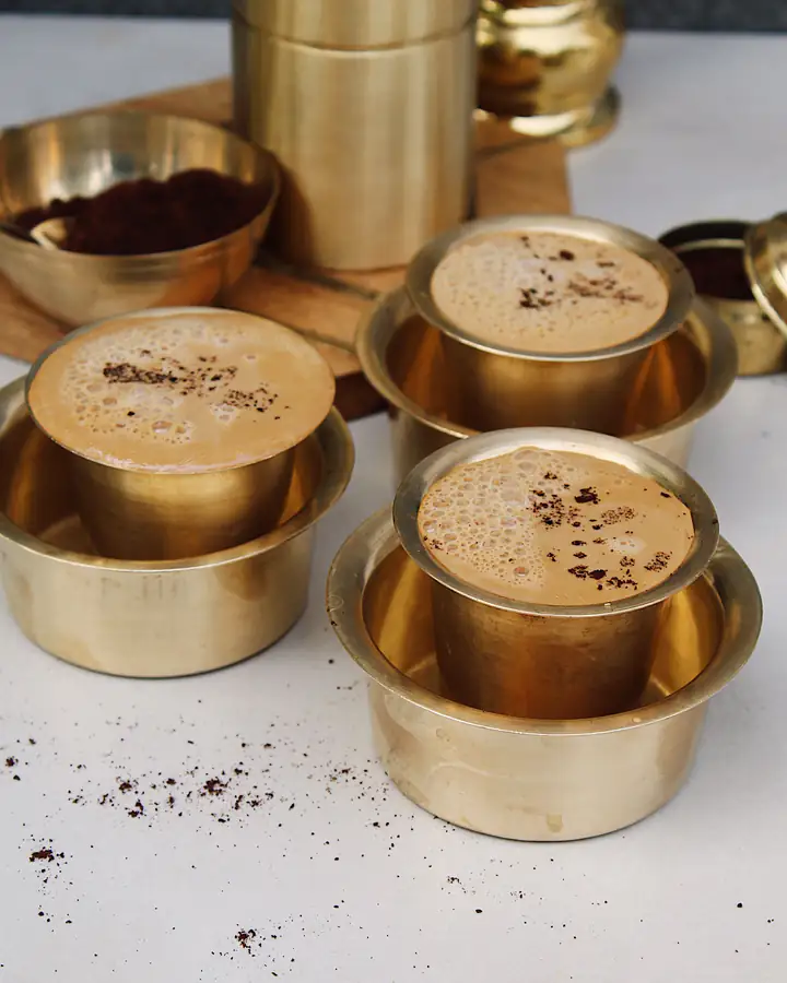 Three brass tumblers of frothy filter coffee set in their davaras, with a brass coffee filter and a bowl of ground coffee behind.